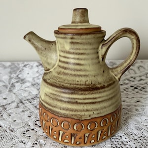 Könnte beinhalten: Keramikkrug mit Deckel, Ausguss und Henkel. Der Krug ist überwiegend beige mit braunen horizontalen Streifen und einem dekorativen Band aus orangefarbenen und braunen Kreisen und Formen in der Nähe des Bodens. Der Griff hat eine Schlaufenform.