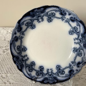 Può includere: Piatto in ceramica bianca con bordo smerlato e design floreale blu. Il bordo del piatto presenta un motivo dettagliato di fiori e viti stilizzate. Il piatto è appoggiato su una tovaglia di pizzo bianco.