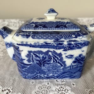 May include: A rectangular ceramic teapot featuring a classic blue and white willow pattern. The design includes detailed illustrations of pagodas, trees, and a bridge. It has a matching lid with a small knob handle, a side spout, and a handle.