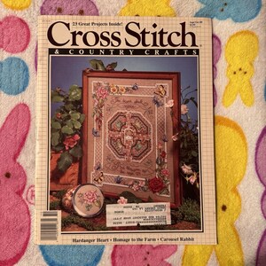 Puede incluir: Una edición vintage de la revista "Cross Stitch & Country Crafts". La portada presenta un diseño de punto de cruz enmarcado con detalles florales y mariposas. También se ve un pequeño recipiente redondo con un diseño floral.