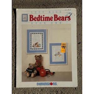 May include: A vintage "Bedtime Bears Book One" craft book by Lucy Rigg. The cover features a cross-stitch design with bears, a moon, and stars. Two framed cross-stitch samplers are displayed, along with two teddy bears and a red ball.
