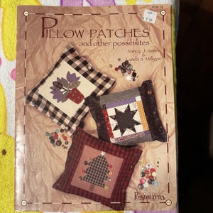 May include: A book cover titled "Pillow Patches and other possibilites" featuring three decorative pillows with various designs. The pillows have floral, star, and Christmas tree patterns. Scattered buttons add detail.