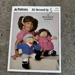 May include: A vintage Patons knitting pattern titled "All Dressed Up" featuring a young girl with two Cabbage Patch Kid dolls. The dolls are wearing knitted outfits in pink and blue. The pattern is in Beehive Astra yarn.
