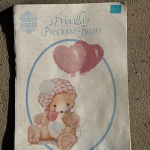 Peut inclure: Un modèle de point de croix intitulé "Priscilla's Precious Bears" représentant un ours en peluche tenant deux ballons roses en forme de cœur. L'ours porte un chapeau et un nœud roses. Le motif est encadré dans un cercle bleu, avec le texte "Designs by Gloria & Pat".
