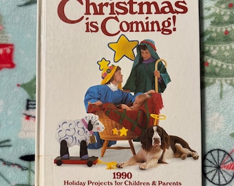 Pode incluir: Um livro vintage intitulado "Christmas is Coming!" com ilustrações de uma cena de presépio, uma ovelha de brinquedo de madeira e um cão. A capa também inclui o ano de 1990 e o subtítulo "Holiday Projects for Children & Parents."