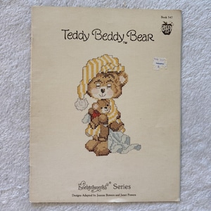 Puede incluir: Una portada de libro vintage con un diseño de punto de cruz de un oso de peluche con gorro de dormir, sosteniendo un oso de peluche más pequeño. El título "Teddy Beddy Bear" se muestra encima de la ilustración. El libro es parte de la "Serie Enchantments".
