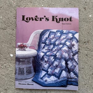 May include: A book cover titled "Lover's Knot, Third Edition" with a pink background. A blue and white quilt is draped over a white wicker chair. A small wicker table holds a floral arrangement. Author: Eleanor Burns.