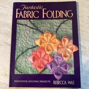 May include: A book titled "Fantastic Fabric Folding" with a purple cover. The cover features four fabric flower designs in orange, peach, and purple hues, set against a green quilted background. The author's name is Rebecca Wat.