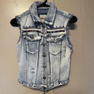 May include: Light blue denim vest with a button-up front, two chest pockets, and decorative trim across the chest. The vest has a distressed look with frayed edges and the brand name "Mud" is visible on the collar.