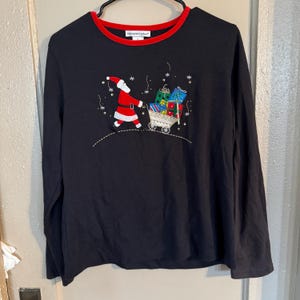 Op de afbeelding: Zwart shirt met lange mouwen en een rode kraag. Het shirt heeft een geborduurd ontwerp van de Kerstman die een kar vol ingepakte cadeaus trekt. Het ontwerp bevat sneeuwvlokken en een stippellijn, wat een feestelijk kerstthema creëert.