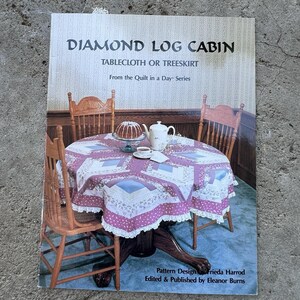 May include: A book cover titled "Diamond Log Cabin" with text about a tablecloth or treeskirt. The image shows a round table set for tea with a patterned tablecloth, cake, and teacups. Wooden chairs surround the table.
