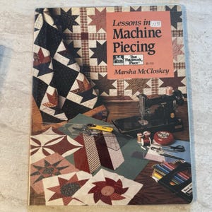 May include: A book titled "Lessons in Machine Piecing" by Marsha McCloskey. The cover features a patchwork quilt design. The image also shows a sewing machine, spools of thread, scissors, and a pincushion, suggesting a quilting theme.