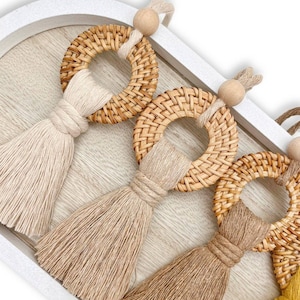 May include: Three decorative ornaments featuring circular woven rattan rings in a light brown tone. Each ring is adorned with a hanging cotton tassel in varying shades of cream, beige, and tan, topped with a small, smooth wooden bead.