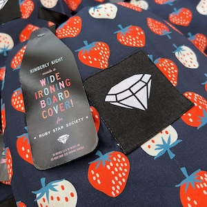 May include: A navy blue ironing board cover with a white diamond logo and a repeating pattern of red and white strawberries. The cover is labeled "Kimberly Kight made a Wide Ironing Board Cover! for Ruby Star Society* 50 inches (L) x 18 inches (W) Its most wide size ironing boards"