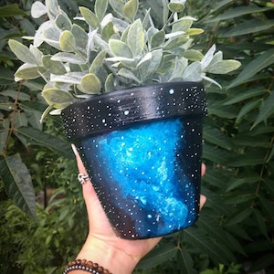 May include: A black terracotta planter with a blue and white galaxy design. The planter is decorated with white dots and has a blue galaxy swirl in the center.