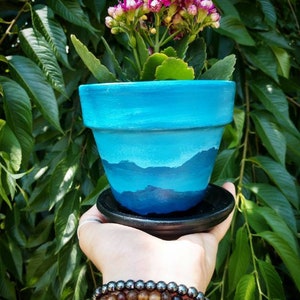 May include: A hand-painted blue ceramic flower pot with a mountain landscape design, holding a flowering plant with pink and white blossoms. The pot sits on a black saucer. The background is a lush green foliage.