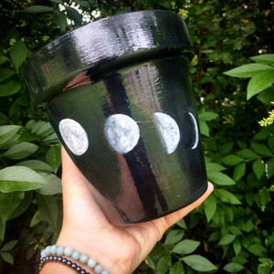 May include: Black terracotta planter with white painted moon phases. The planter is painted with a full moon, a half moon, a crescent moon, and a new moon.