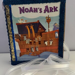 May include: A children's book titled "Noah's Ark" with a colorful illustration of the ark, animals, and text. The book is framed with a blue crocheted border and a white ribbon.