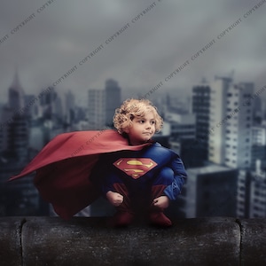 Może przedstawiać: Dziecko ubrane w strój Supermana, w niebiesko-czerwonym kostiumie z kultowym emblematem "S". Dziecko kuca na gzymsie z panoramą miasta w tle, pod zachmurzonym niebem. Czerwona peleryna powiewa na wietrze.