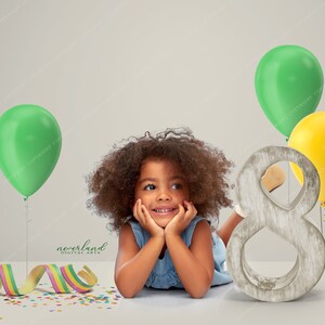 May include: A young child smiles, resting on their arms, with green and yellow balloons and a wooden number eight. Colorful confetti and a ribbon add to the festive birthday theme. The image is from Neverland Digital Arts.