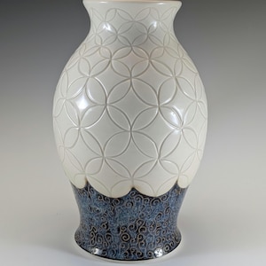 Può includere: Un vaso di ceramica bianco con un motivo. La parte superiore presenta un motivo a cerchi ripetuti, mentre la base ha un design a spirale blu e marrone. Il vaso ha una parte superiore svasata e una finitura lucida.