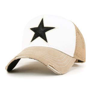 May include: A white and tan trucker hat with a black star applique. The hat features a mesh back, a distressed brim, and a white front panel with a black star. The brim is tan, and the hat has a classic trucker style.