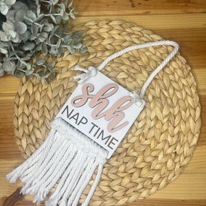 Pode incluir: Uma placa de madeira branca com as palavras "shh NAP TIME" em letras rosa e brancas. A placa é adornada com uma franja de macramê branca e uma corda branca para pendurar. A placa é colocada sobre um sousplat tecido.