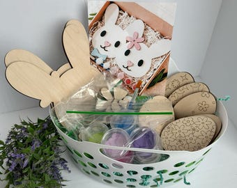 以下が含まれることがあります： イースターのクラフトプロジェクトのためのクラフト用品が入った白いバスケット。バスケットには、木製のウサギの耳、木製の卵、絵の具、キラキラが入っています。