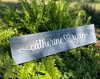 May include: A rustic wooden sign with the names "Catherine" and "Ryan" written in white cursive lettering with a heart symbol between the names.