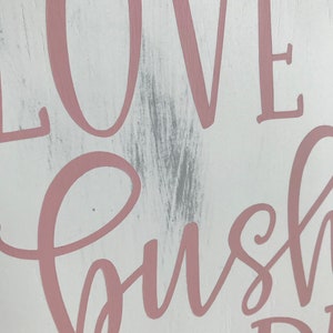 May include: A white wooden sign with the words "Love, bush, and dreams" written in pink cursive lettering.