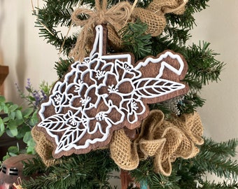 May include: A wooden ornament shaped like the state of West Virginia, decorated with white floral designs. The ornament is hanging on a green Christmas tree with burlap ribbon.