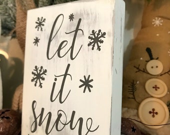 Könnte beinhalten: Ein weißes Holzschild mit dem Text "Let it snow" in schwarzer Kursivschrift. Das Schild ist mit kleinen Schneeflocken verziert.