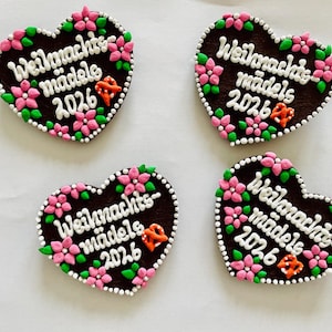 Könnte beinhalten: Vier herzförmige, dunkelbraune Lebkuchen mit weißer Zuckerschrift "Weihnachts-mädels 2026". Jeder Lebkuchen ist mit rosafarbenen Zuckerblüten, grünen Blättern und einer kleinen orangefarbenen Brezel verziert, umrandet von weißen Zuckerperlen.