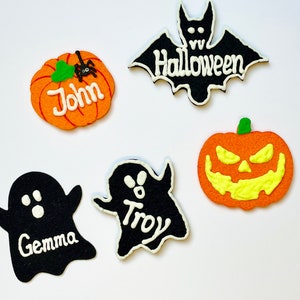 Könnte beinhalten: Fünf Halloween-Kekse, die mit Zuckerguss verziert sind. Zwei Kekse sind in Form von Geistern, einer mit dem Namen "Gemma" und der andere mit dem Namen "Troy". Ein Keks ist in Form eines Kürbisses mit einem gruseligen Gesicht. Ein Keks ist in Form eines Kürbisses mit dem Namen "John" und einer kleinen Spinne oben drauf. Ein Keks ist in Form einer Fledermaus mit dem Wort "Halloween" darauf.