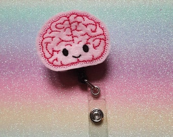 May include: A pink felt badge reel featuring a stitched brain design with a smiling face. The brain has dark pink thread detailing for the cerebral cortex and two black eyes. A clear plastic strap with a metal snap button is attached below.