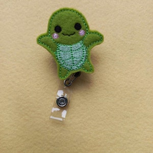 May include: A green felt turtle badge reel featuring embroidered details, including black eyes, pink cheeks, and a light green shell pattern. The turtle is attached to a clear plastic strap with a metal snap button and a retractable cord mechanism.