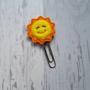 Peut inclure: Un trombone en métal argenté surmonté d'un ornement en feutrine jaune en forme de soleil. Le soleil présente une bordure orange cousue, un visage circulaire jaune vif, ainsi que des yeux fermés et une bouche souriante brodés en noir.