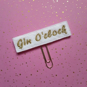 May include: A white rectangular felt bookmark featuring the embroidered gold text Gin O'clock, attached to a silver metal paperclip. The felt piece has a stitched border and is positioned horizontally with the paperclip extending from the bottom.