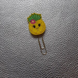 Peut inclure: Un trombone en métal surmonté d'un ananas en feutrine jaune avec des feuilles vertes brodées, une petite fleur rose et un visage souriant aux yeux noirs. L'ananas présente une texture quadrillée cousue avec un fil de couleur dorée.