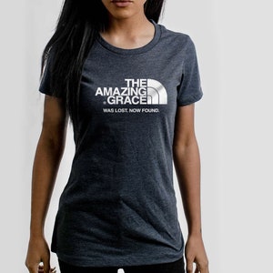 Puede incluir: Una camiseta gris oscuro jaspeada con un estampado gr&aacute;fico blanco de "The Amazing Grace" con un logotipo de pico de monta&ntilde;a. El texto debajo del logotipo dice "Was lost. Now found."