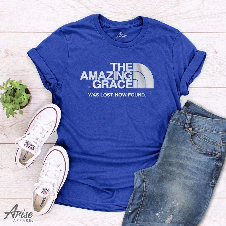 Puede incluir: Una camiseta azul real con un dise&ntilde;o gr&aacute;fico blanco que dice "The Amazing Grace Was Lost, Now Found." El dise&ntilde;o gr&aacute;fico es una parodia del logotipo de una marca de exteriores popular.