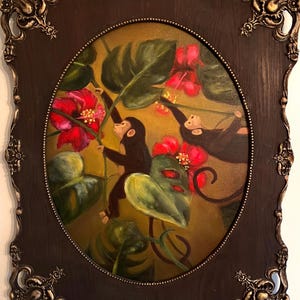 May include: An oval painting of two monkeys reaching for red flowers and green leaves. The artwork is framed in a dark brown, ornate frame with gold accents. The background is a warm, golden color.
