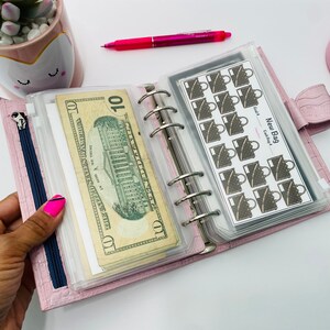 May include: A pink, leather-like planner with a zipper and silver rings, holding a ten-dollar bill and stickers. The stickers have a purse design and the text "New Bag". A pink pen and calculator are also visible.