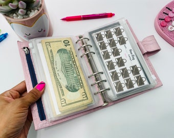 May include: A pink, leather-like planner with a zipper and silver rings, holding a ten-dollar bill and stickers. The stickers have a purse design and the text "New Bag". A pink pen and calculator are also visible.