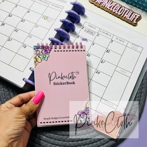May include: A pink Pinkecloth StickerBook with floral accents and the text "Simple Budget Essentials" is held next to a calendar. The calendar has a purple spiral binding and a wooden sign that says "Pinkeclothlife".
