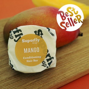 May include: A round, white, paper-wrapped bar of mango-scented conditioning hair soap. The label reads "Superfly Soap" and "Mango Conditioning Hair Bar". The bar is sitting on a wooden surface with a mango fruit in the background. A white circle with the text "Best Seller" is partially visible in the top right corner of the image.