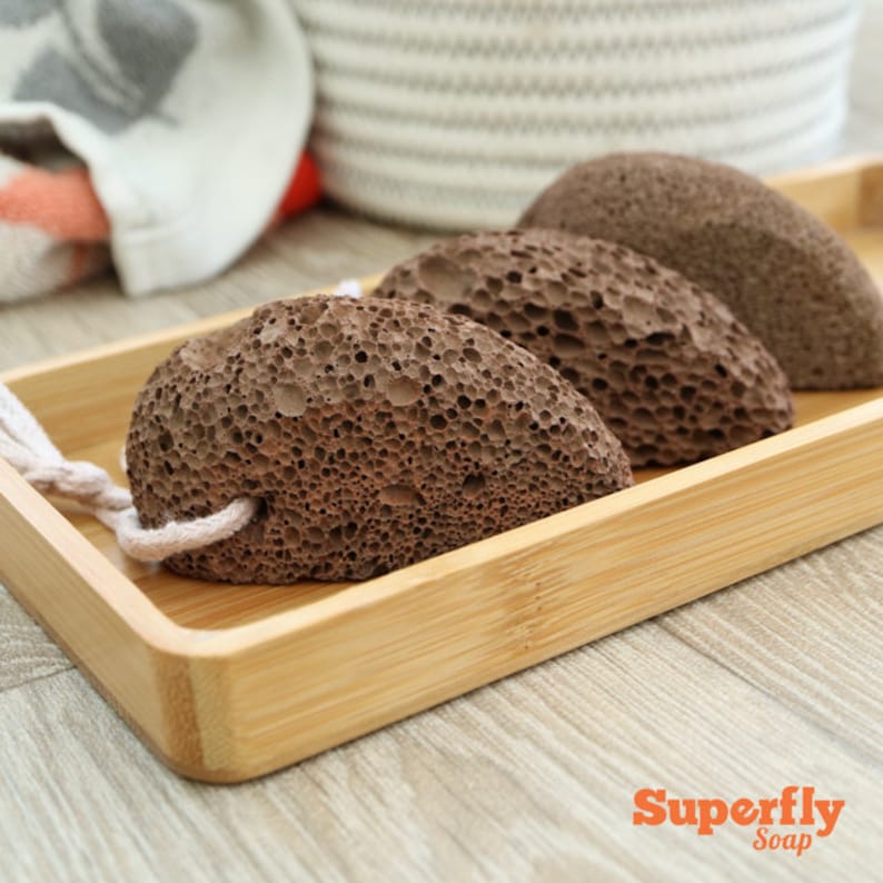 May include: Three brown pumice stones in a wooden tray. The stones are rough and have a textured surface. They are used for removing calluses and dead skin from the feet.