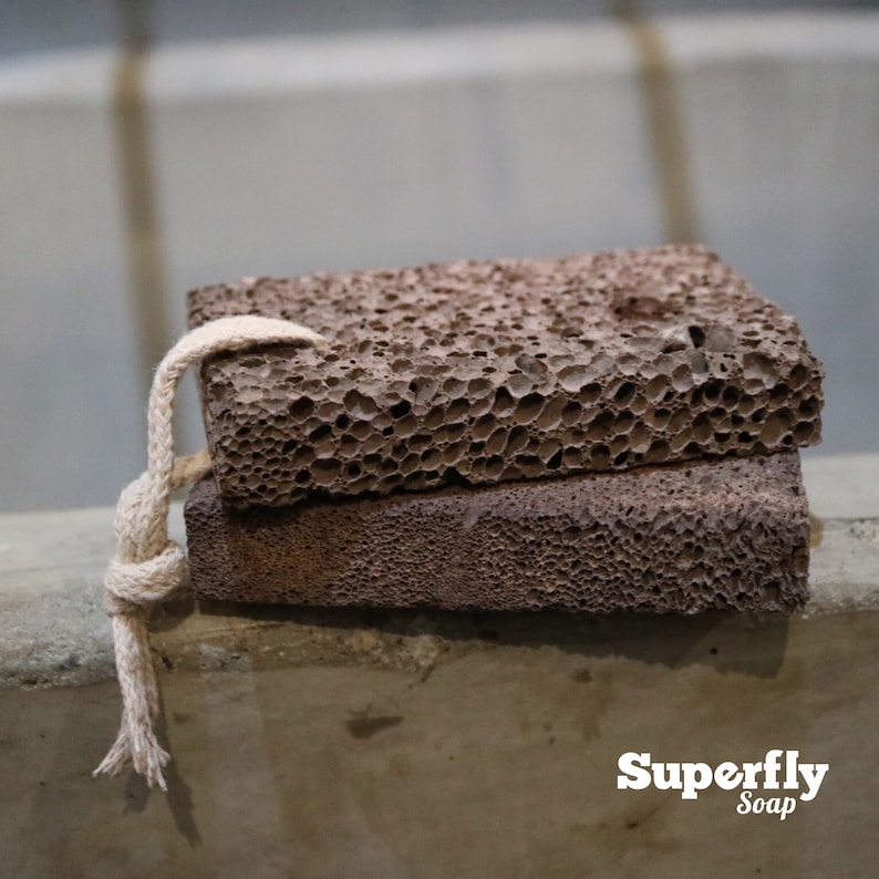 May include: Two brown pumice stones stacked on top of each other. A beige rope is tied around the top stone. The stones are sitting on a gray concrete surface. Superfly Soap is printed in white text on the bottom right corner of the image.