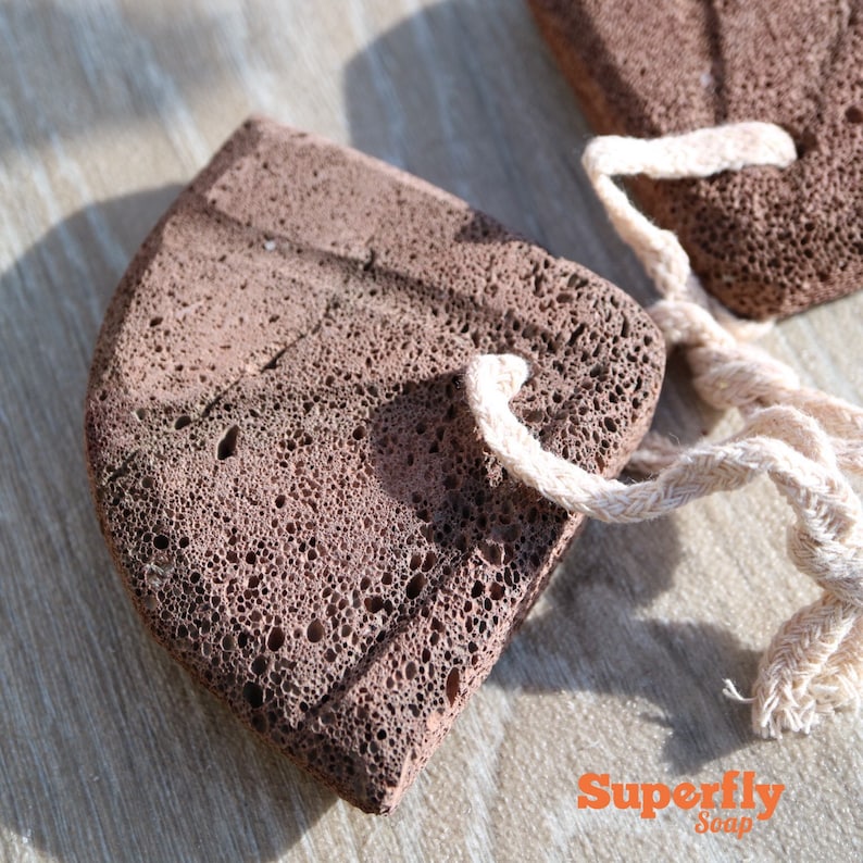May include: Two brown pumice stones with a white string attached to each. The stones are rough and have a porous texture. The stones are on a wooden surface.