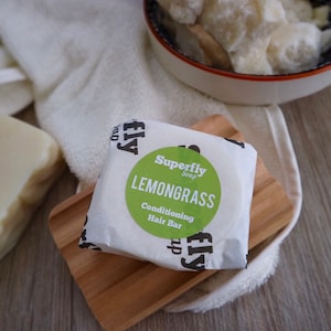 Op de afbeelding: Een witte zeepstaaf verpakt in wit papier met een groene cirkel waarop "Superfly Soap Lemongrass Conditioning Hair Bar" staat.